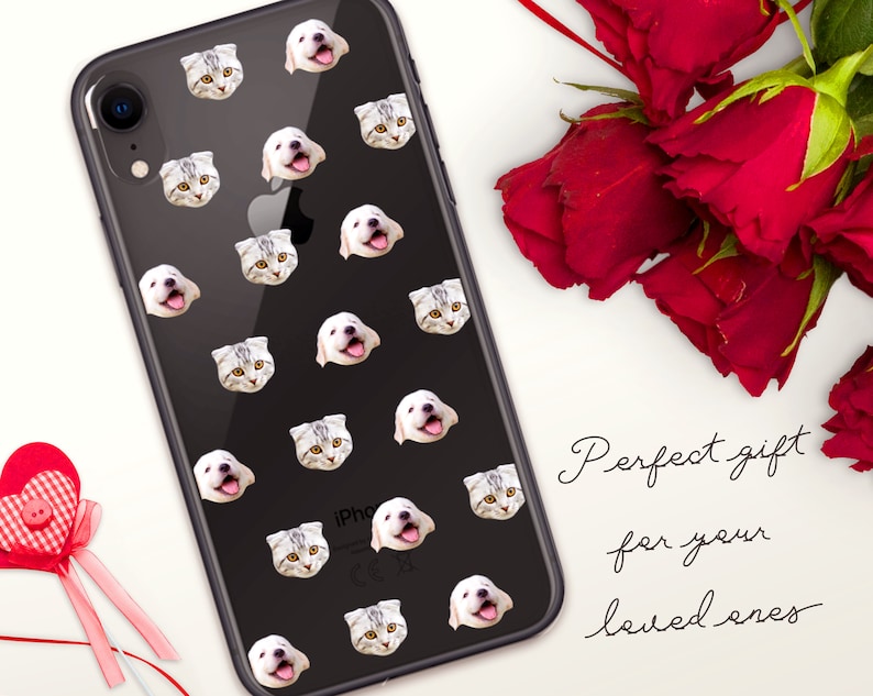 May include: A black iPhone case with a repeating pattern of white cat and dog faces. The case is on a white surface with a red rose and a red heart.