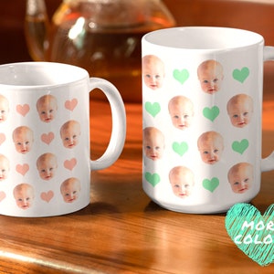 May include: Two white ceramic mugs with a repeating pattern of baby faces and hearts. One mug has pink hearts and the other has green hearts. The text "MORE COLORS" is written in a green heart.