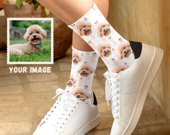 May include: White socks with a repeating pattern of a dog's face. The dog is a small, light brown poodle with a happy expression. The socks are worn on a person's feet with white sneakers.