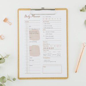 Op de afbeelding: Een dagelijkse planner met een bruin clipboard en een gouden pen. De planner heeft een schema, een to-dolijst en secties voor het bijhouden van waterinname, maaltijden en dankbaarheid. De tekst op de planner zegt "Daily Planner" en "Wed. 04 of July".
