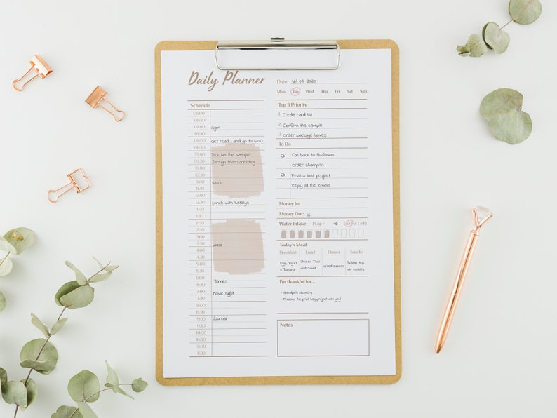 Op de afbeelding: Een dagelijkse planner met een bruin clipboard en een gouden pen. De planner heeft een schema, een to-dolijst en secties voor het bijhouden van waterinname, maaltijden en dankbaarheid. De tekst op de planner zegt "Daily Planner" en "Wed. 04 of July".