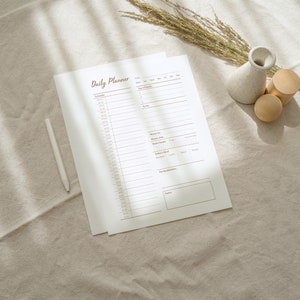 Op de afbeelding: Een witte dagelijkse planner met de bruine titel "Daily Planner" op een beige linnen achtergrond. Er zijn twee houten cilinders en een witte keramische vaas met gedroogd gras.