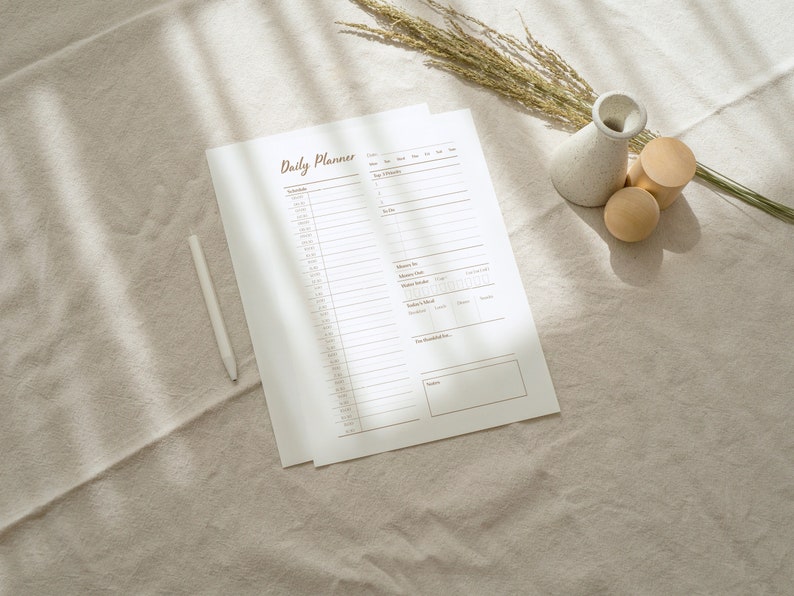 Op de afbeelding: Een witte dagelijkse planner met de bruine titel "Daily Planner" op een beige linnen achtergrond. Er zijn twee houten cilinders en een witte keramische vaas met gedroogd gras.