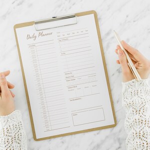 Op de afbeelding: Een dagelijkse planner op een klembord met een zilveren pen. De planner heeft secties voor schema, top 3 prioriteiten, te doen, geheugenbox, waterinname, gezonde gewoonten en een to-do-checklist.