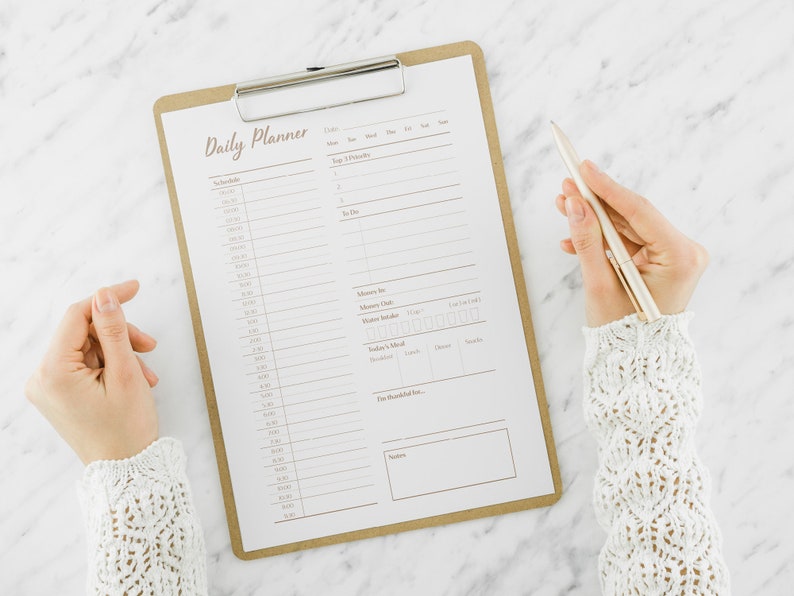 Op de afbeelding: Een dagelijkse planner op een klembord met een zilveren pen. De planner heeft secties voor schema, top 3 prioriteiten, te doen, geheugenbox, waterinname, gezonde gewoonten en een to-do-checklist.