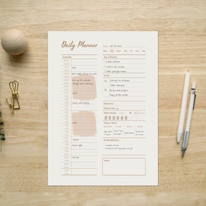 Op de afbeelding: Een dagelijkse planner met een witte achtergrond en bruine tekst. De planner bevat secties voor planning, topprioriteiten, geld in, geld uit, waterinname, maaltijd van vandaag en dankbaarheid. De planner ligt op een houten oppervlak.