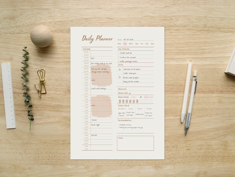 Op de afbeelding: Een dagelijkse planner met een witte achtergrond en bruine tekst. De planner bevat secties voor planning, topprioriteiten, geld in, geld uit, waterinname, maaltijd van vandaag en dankbaarheid. De planner ligt op een houten oppervlak.