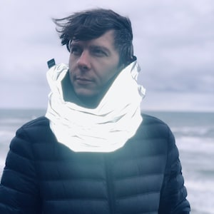 Puede incluir: Un hombre con una chaqueta acolchada azul oscuro y una bufanda plateada reflectante alrededor del cuello. La bufanda está doblada y tiene un acabado brillante e iridiscente.