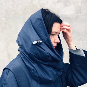 May include: A navy blue hooded coat. The coat has a large hood that is worn over the head. The coat is made of a smooth, possibly water-resistant material. A small tag with a circular design is visible on the hood.