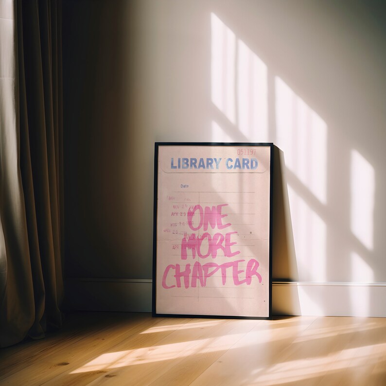 K&ouml;nnte beinhalten: Ein gerahmtes rosa und wei&szlig;es Poster mit dem Text "LIBRARY CARD" und "ONE MORE CHAPTER" in rosa Schrift. Das Poster liegt auf einem Holzboden mit einem braunen Vorhang im Hintergrund.