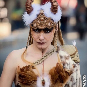 May include: A woman wearing a costume with a white feathered headdress, gold and brown horns, and a gold and brown beaded mask. She is also wearing a brown feather bra and a brown leather strap across her chest.