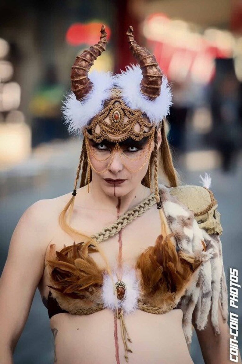 May include: A woman wearing a costume with a white feathered headdress, gold and brown horns, and a gold and brown beaded mask. She is also wearing a brown feather bra and a brown leather strap across her chest.