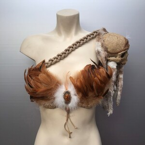 May include: A brown and white feather bra with a skull and rope shoulder piece. The bra has a brown stone embellishment in the center.