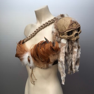 May include: A brown and white feathered costume piece with a skull and fur accents. The skull is decorated with a gold-colored band.