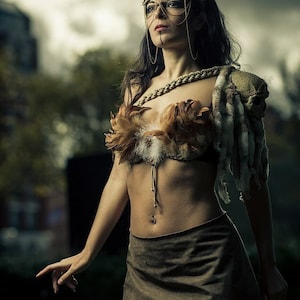 May include: A woman wearing a brown leather skirt and a feathered bra with a gold chain necklace. She is wearing a headdress with horns and a gold chain with a pendant. The background is out of focus.