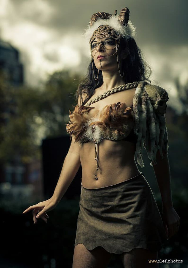 May include: A woman wearing a brown leather skirt and a feathered bra with a gold chain necklace. She is wearing a headdress with horns and a gold chain with a pendant. The background is out of focus.
