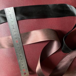 May include: Two satin ribbons, one black and one brown, are displayed against a burgundy background. A metal ruler shows measurements in inches. The ribbons are approximately 1 inch wide.