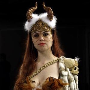 May include: A woman wearing a gold and white headdress with horns, a gold chainmail mask, and a brown and white feathered costume.