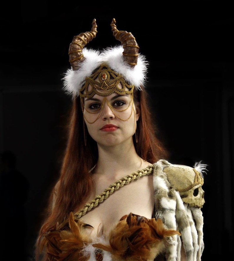 May include: A woman wearing a gold and white headdress with horns, a gold chainmail mask, and a brown and white feathered costume.