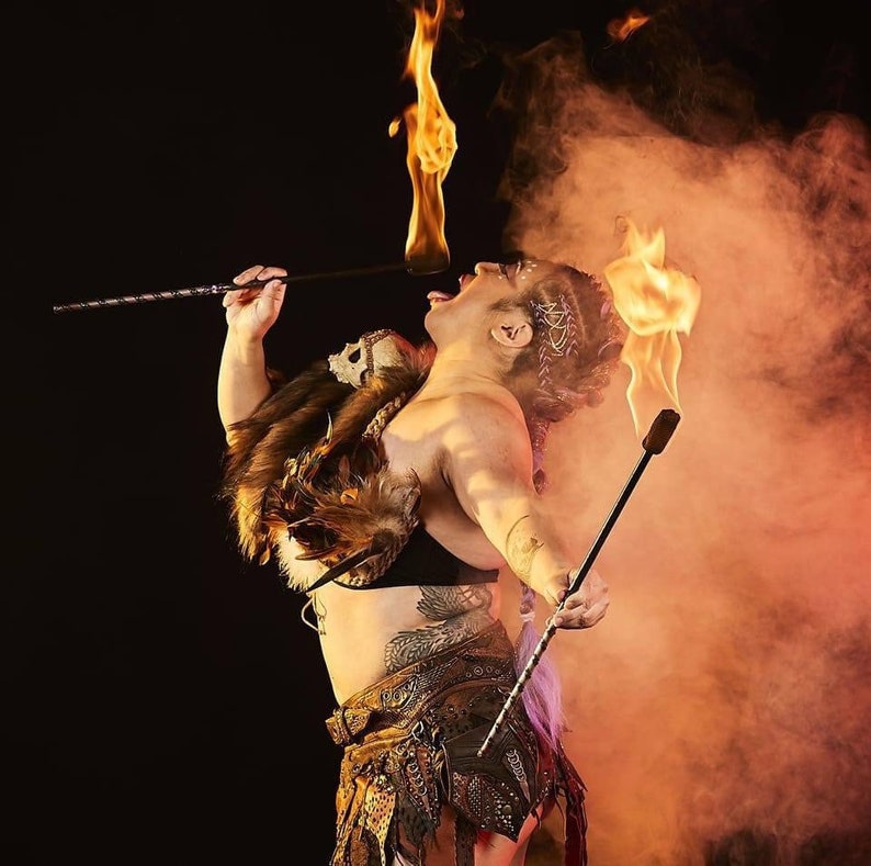 May include: A person wearing a brown leather and metal costume with feathers and a skull detail, performing a fire breathing act with a long stick. The person is surrounded by smoke and fire.