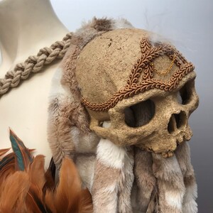 May include: A faux skull with a gold chain and brown fur trim. The skull is decorated with a brown, leafy headband. The skull is sitting on a brown and white fur base.