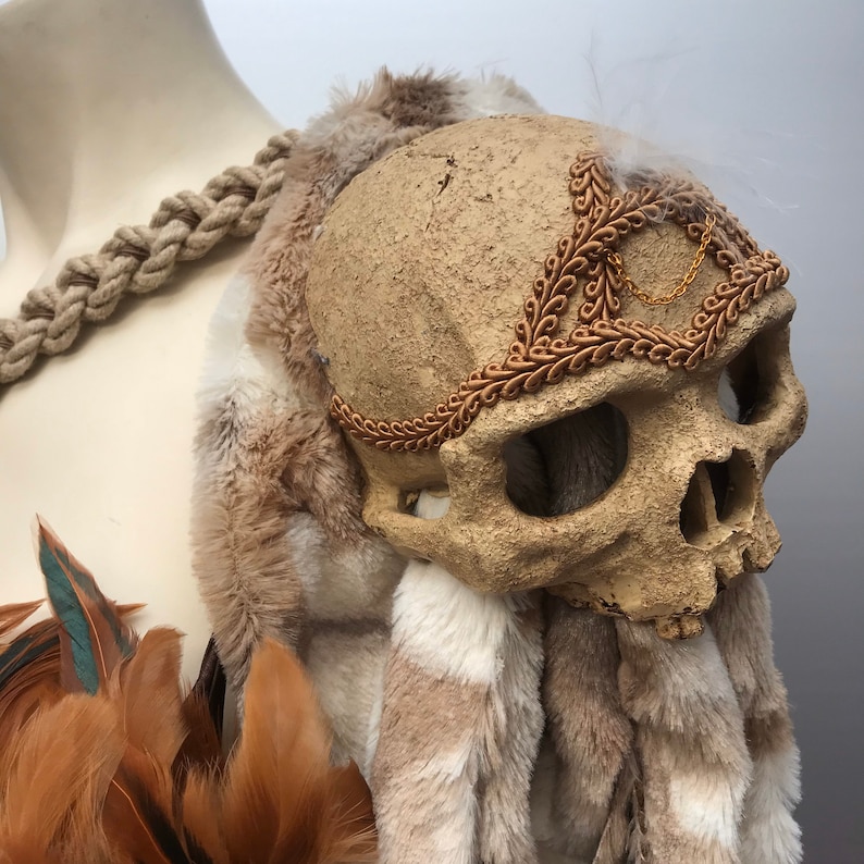May include: A faux skull with a gold chain and brown fur trim. The skull is decorated with a brown, leafy headband. The skull is sitting on a brown and white fur base.