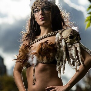 May include: A woman wearing a costume with a skull shoulder piece, a feathered bra, and a gold headpiece with horns. She is standing with her hands on her hips and looking up.