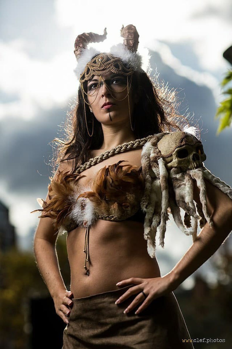 May include: A woman wearing a costume with a skull shoulder piece, a feathered bra, and a gold headpiece with horns. She is standing with her hands on her hips and looking up.