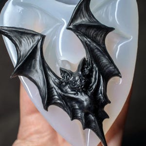 May include: A silicone mold for making a black bat figurine. The mold is clear and the bat is in a flying position with its wings spread wide.