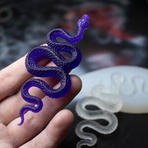 May include: A purple resin snake figurine with a coiled body and a raised head. The snake is being held in a hand, and a white silicone mold of a snake is visible in the background.
