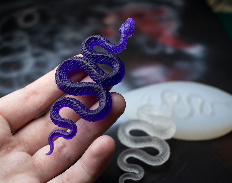 May include: A purple resin snake figurine with a coiled body and a raised head. The snake is being held in a hand, and a white silicone mold of a snake is visible in the background.