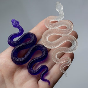May include: Two resin snake sculptures, one in a clear translucent color and the other in a deep purple color. Both snakes are coiled in a similar pose.