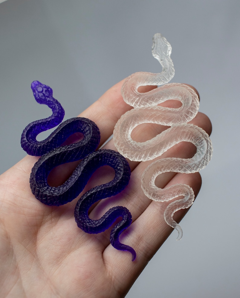 May include: Two resin snake sculptures, one in a clear translucent color and the other in a deep purple color. Both snakes are coiled in a similar pose.