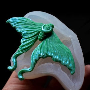 Puede incluir: Una mariposa de resina verde con un dije de luna creciente, en un molde de silicona transparente. La mariposa tiene alas detalladas y una cola rizada.