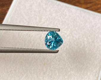 May include: A faceted, heart-shaped, bright teal gemstone is held by silver tweezers. The gem is set against a white background, with a wooden surface visible in the background. The gemstone's facets reflect light, creating a sparkling effect.