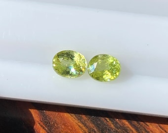 May include: Two oval-shaped green gemstones, likely peridot, on a white surface. The stones are translucent and have a slight shimmer.
