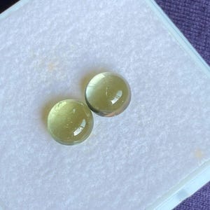 May include: Two round, light green gemstones with a polished, reflective surface. The stones are set against a white background inside a square container. The gemstones have a translucent quality.