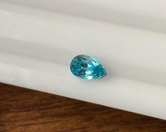 May include: A pear-shaped, faceted gemstone in shades of turquoise and aqua blue. The gem is set against a white background with a wooden surface below. The gemstone's cut allows light to reflect, creating a sparkling effect.