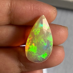 May include: A faceted, pear-shaped opal gemstone. The gem exhibits a vibrant play-of-colour, with flashes of green, yellow, and blue. The stone is held between fingers, showcasing its polished surface and internal iridescence. The opal's cut highlights its natural beauty.