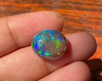 May include: A round, iridescent opal gemstone with a dark grey base and flashes of green, blue, and purple.