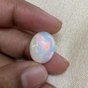 Puede incluir: Una piedra preciosa de ópalo ovalada blanca con colores arcoíris iridiscentes.