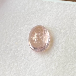 Puede incluir: Una gema ovalada y translúcida de un suave tono rosado. La superficie pulida refleja la luz, mostrando patrones internos. La gema se encuentra sobre un fondo blanco, resaltando su color y claridad. Adecuada para la creación de joyas.