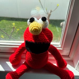 Może przedstawiać: Czerwona pluszowa zabawka wykonana na szydełku z czarnymi oczami, żółtym nosem i czarnymi ustami. Zabawka siedzi na białym parapecie.