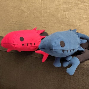 以下が含まれることがあります： 赤いぬいぐるみと青いぬいぐるみの2つのぬいぐるみ。黒い目と歯があります。ぬいぐるみは茶色のソファの上に置かれています。