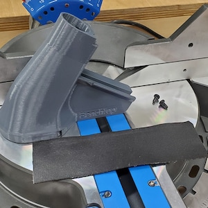 May include: A gray 3D printed plastic attachment for a miter saw. The attachment has the word "Bandshop" printed on it. The attachment is sitting on a miter saw with a black fabric strap.