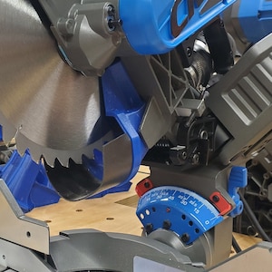 May include: A close-up of a blue and gray miter saw with a silver blade. The saw has a blue and red protractor with markings from 0 to 45 degrees. The saw is sitting on a wooden surface.