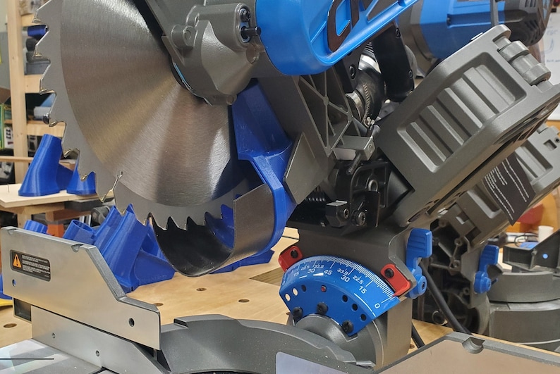 May include: A close-up of a blue and gray miter saw with a silver blade. The saw has a blue and red protractor with markings from 0 to 45 degrees. The saw is sitting on a wooden surface.