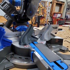 May include: A close-up of a miter saw with two gray plastic pieces attached to the saw. The saw is on a wooden table with a black cord and a pair of scissors.