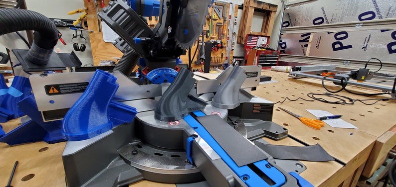 May include: A close-up of a miter saw with two gray plastic pieces attached to the saw. The saw is on a wooden table with a black cord and a pair of scissors.