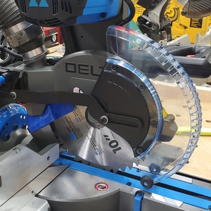 May include: A close-up of a black and blue Delta power miter saw with a clear plastic blade guard. The saw is on a wooden table and the blade is visible.